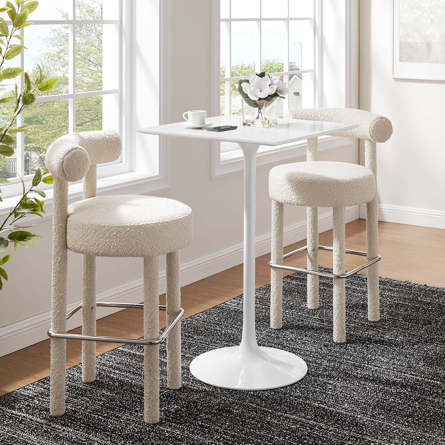 White tulip pedestal table with two boucle upholstered bar stools and chrome foot rails in a sunlit breakfast nook.