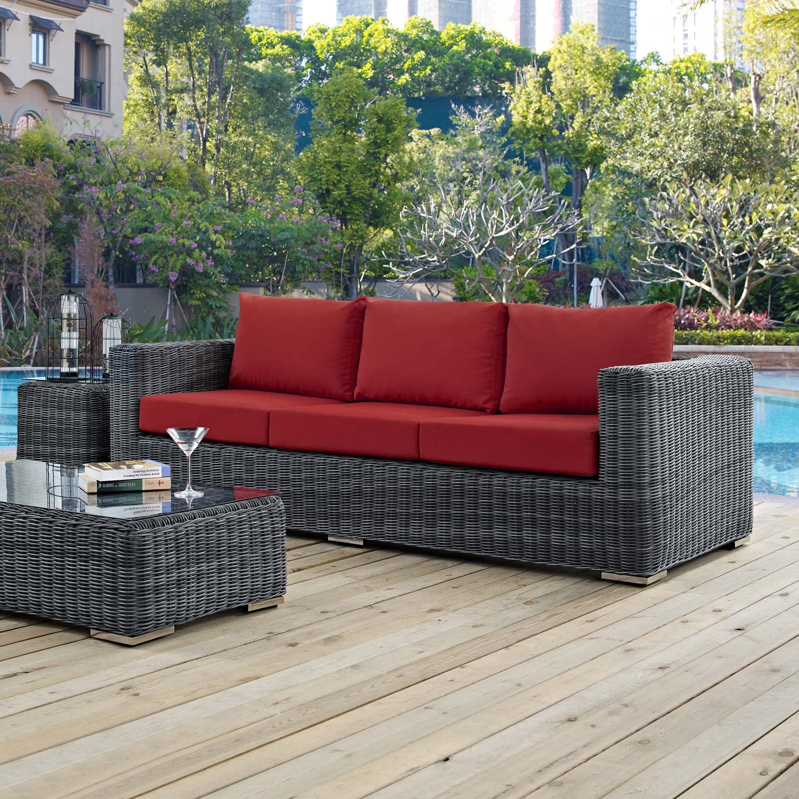 Grey wicker outdoor sofa with red cushions and glass-top coffee table holding books and a cocktail.