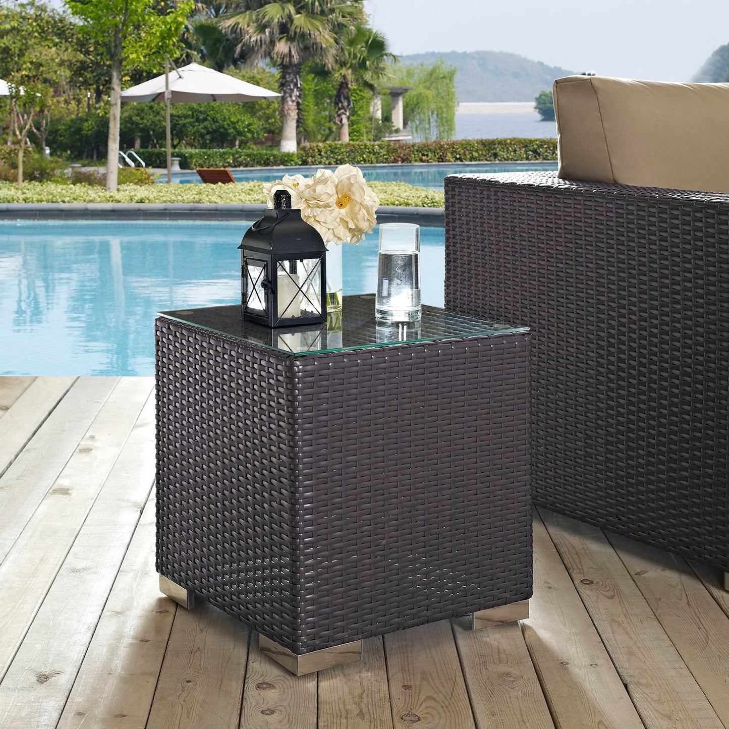Dark brown wicker square outdoor side table with glass top beside a pool; lantern and flower decor.