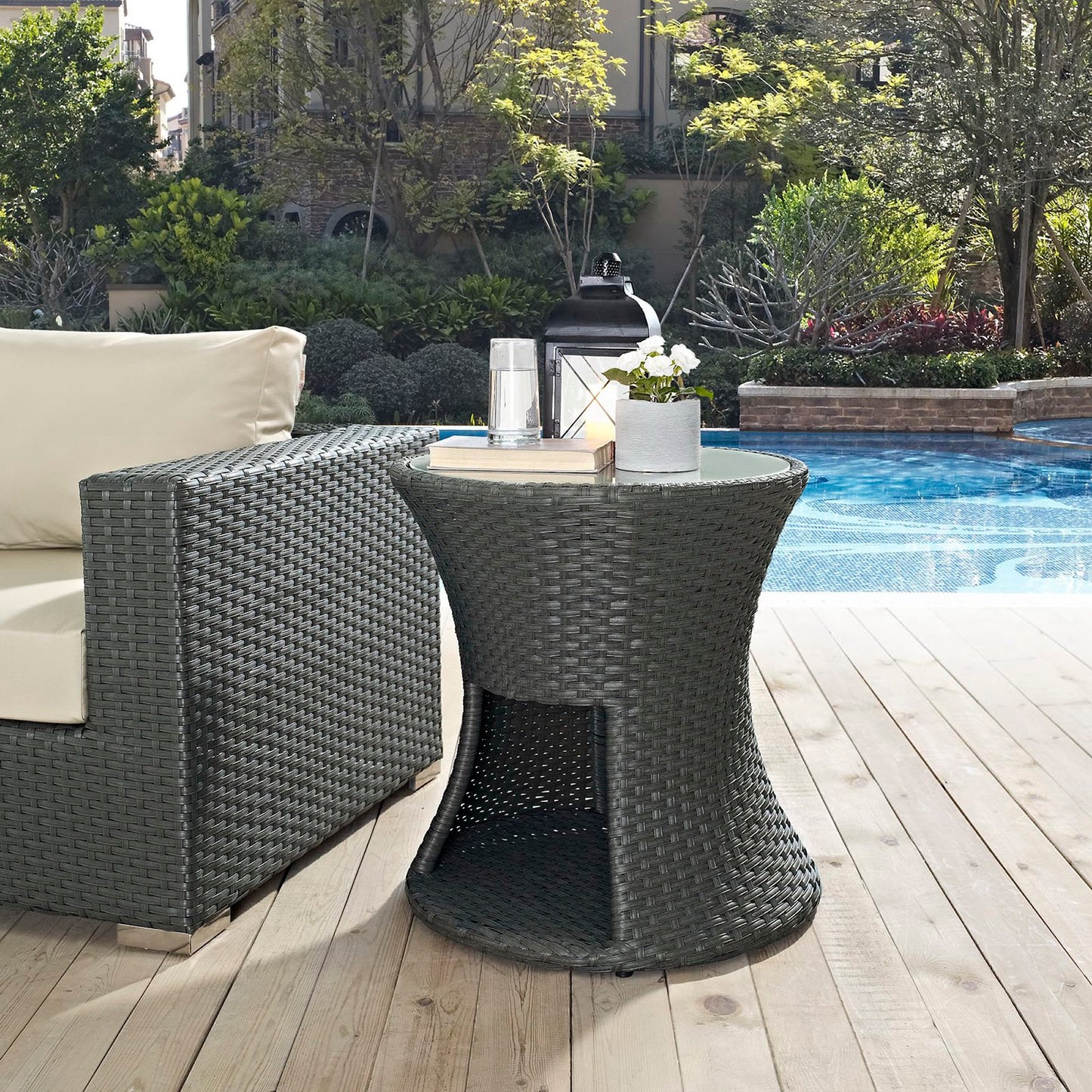 Chocolate-colored round outdoor patio side table with a lower shelf, woven wicker, beside a beige cushioned outdoor sofa poolside on a wooden deck.