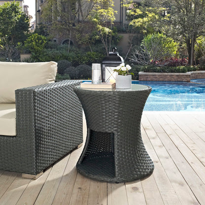 Chocolate-colored round outdoor patio side table with a lower shelf, woven wicker, beside a beige cushioned outdoor sofa poolside on a wooden deck.