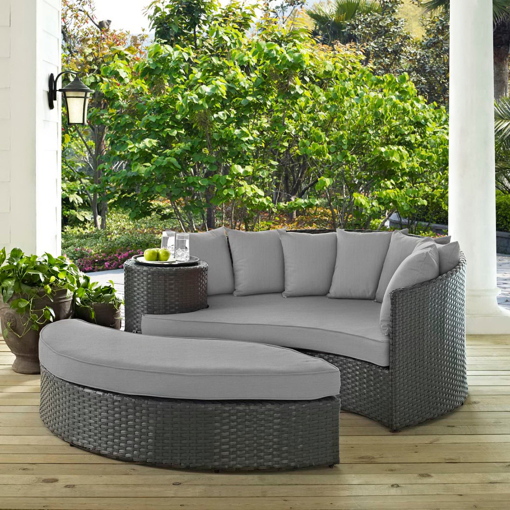Outdoor canvas gray wicker daybed with light gray cushions and a glass-top table amid lush greenery.
