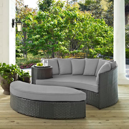 Outdoor canvas gray wicker daybed with light gray cushions and a glass-top table amid lush greenery.