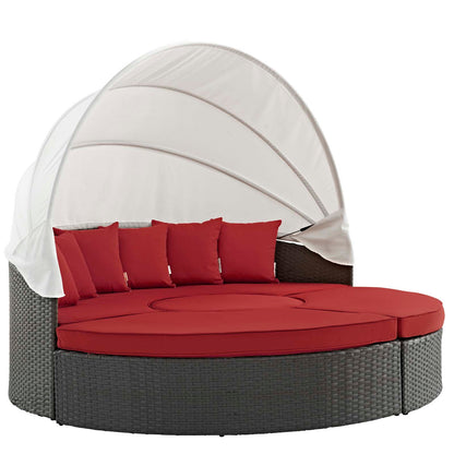 Round outdoor wicker sectional daybed with red cushions and pillows under a curved white canopy.