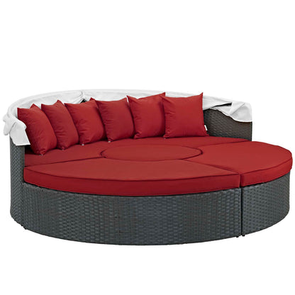 Round gray wicker outdoor daybed with red cushions and pillows, white throw blanket draped over the back.