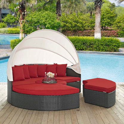 Poolside circular gray wicker outdoor daybed with red cushions, beige canopy, and matching ottoman