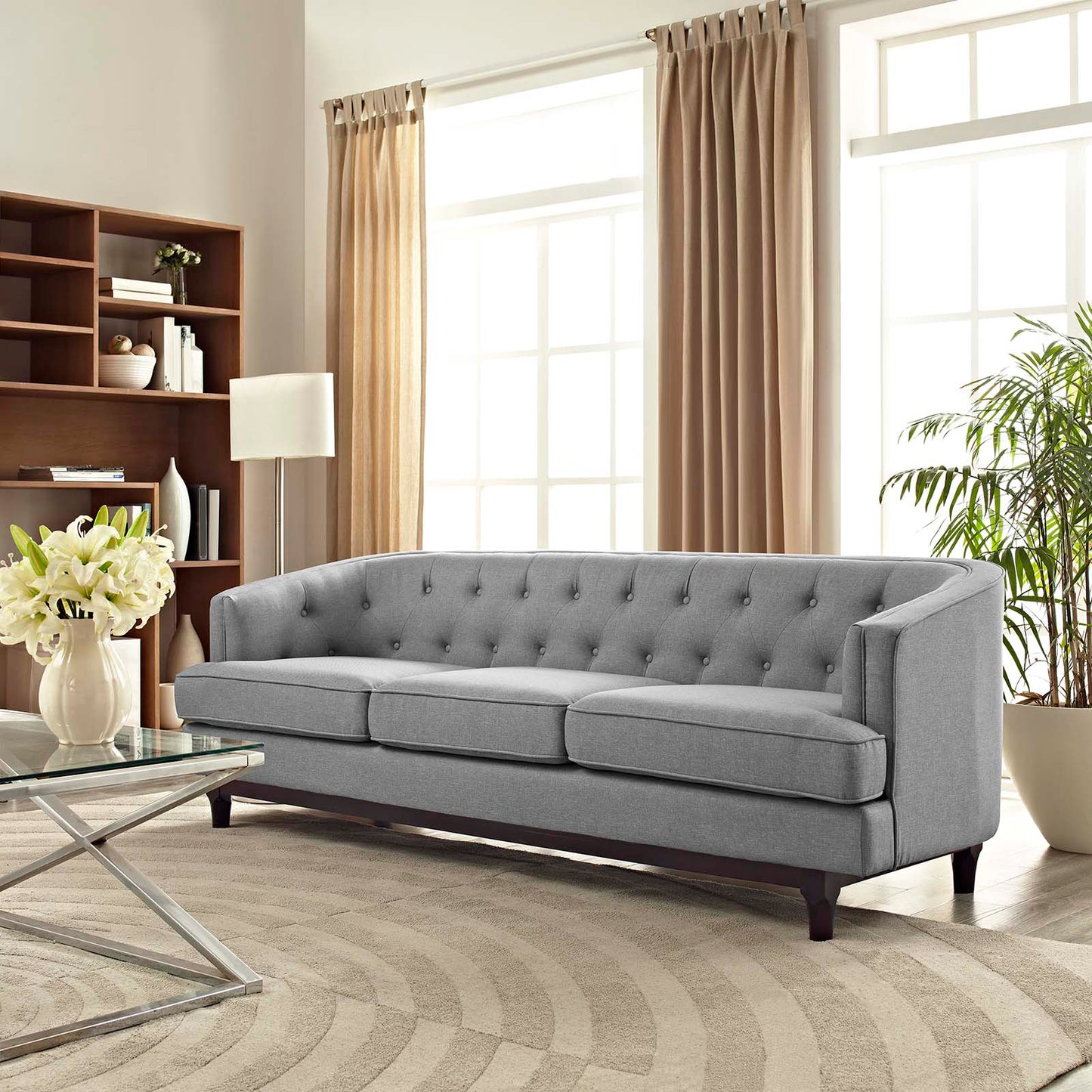 Light gray tufted sofa with square arms and two cushions in a bright living room with beige curtains and glass coffee table.