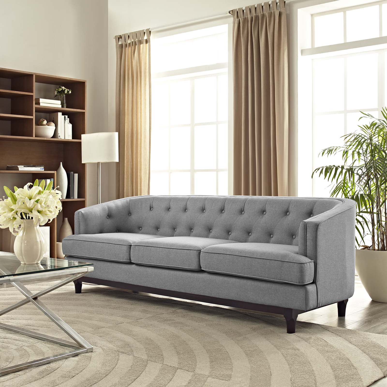 Light gray tufted sofa with square arms and two cushions in a bright living room with beige curtains and glass coffee table.