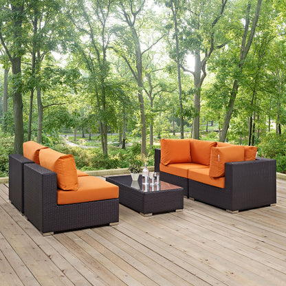 Espresso wicker outdoor sectional set with orange cushions and a glass coffee table on a wooden deck amid lush trees.