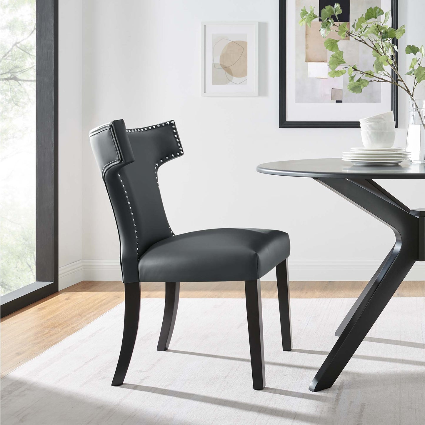 Dark gray vegan-leather dining chair with nailhead trim and curved back, black wood legs beside a round glass-top table.
