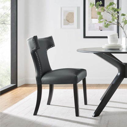 Dark gray vegan-leather dining chair with nailhead trim and curved back, black wood legs beside a round glass-top table.