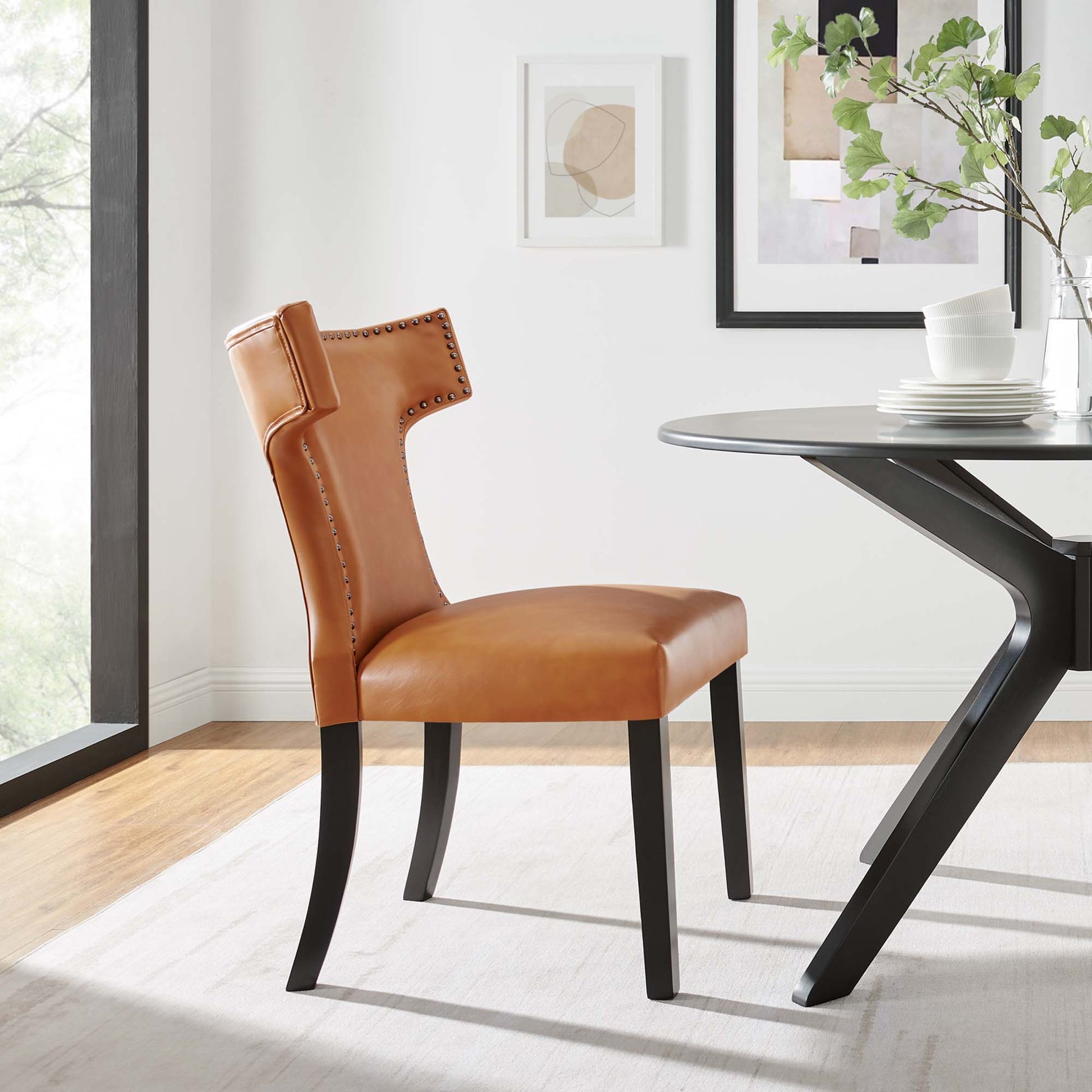 Tan vegan leather dining chair with nailhead trim, curved high back, and black tapered legs.