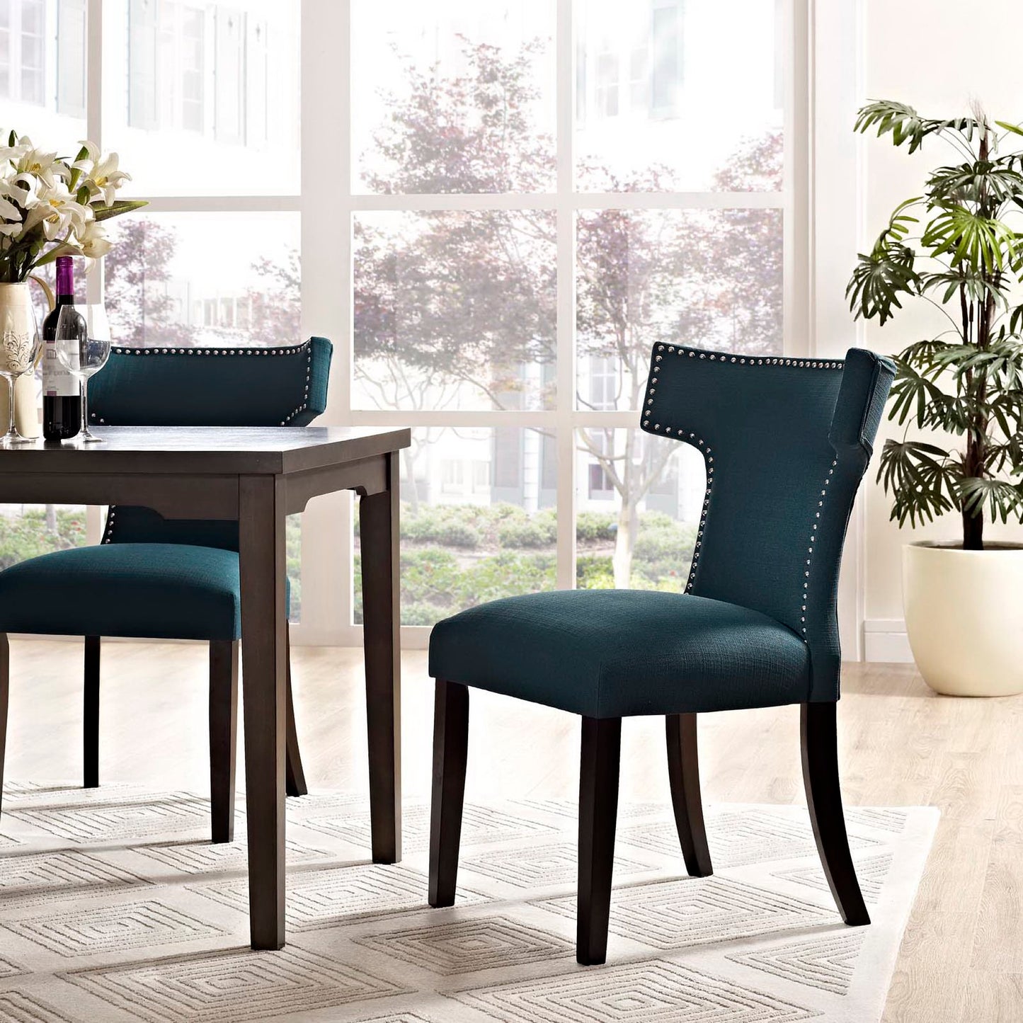 Blue fabric dining chair with a curved back and nailhead trim beside a dark wood table in a bright dining area.
