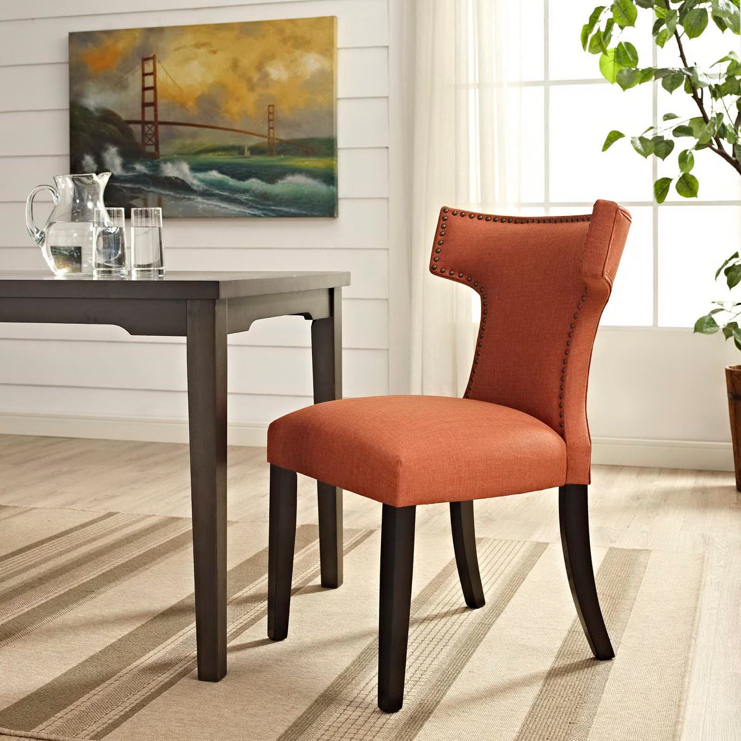 Orange upholstered dining chair with curved back and nailhead trim, dark wood legs beside a table in a bright dining room.