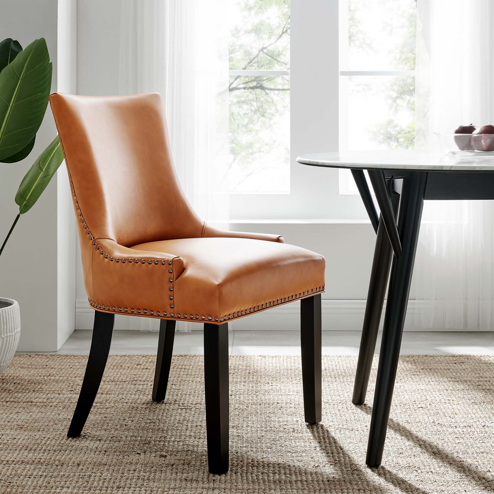 Tan leather dining chair with nailhead trim, curved back, and dark wooden legs beside a round white table.