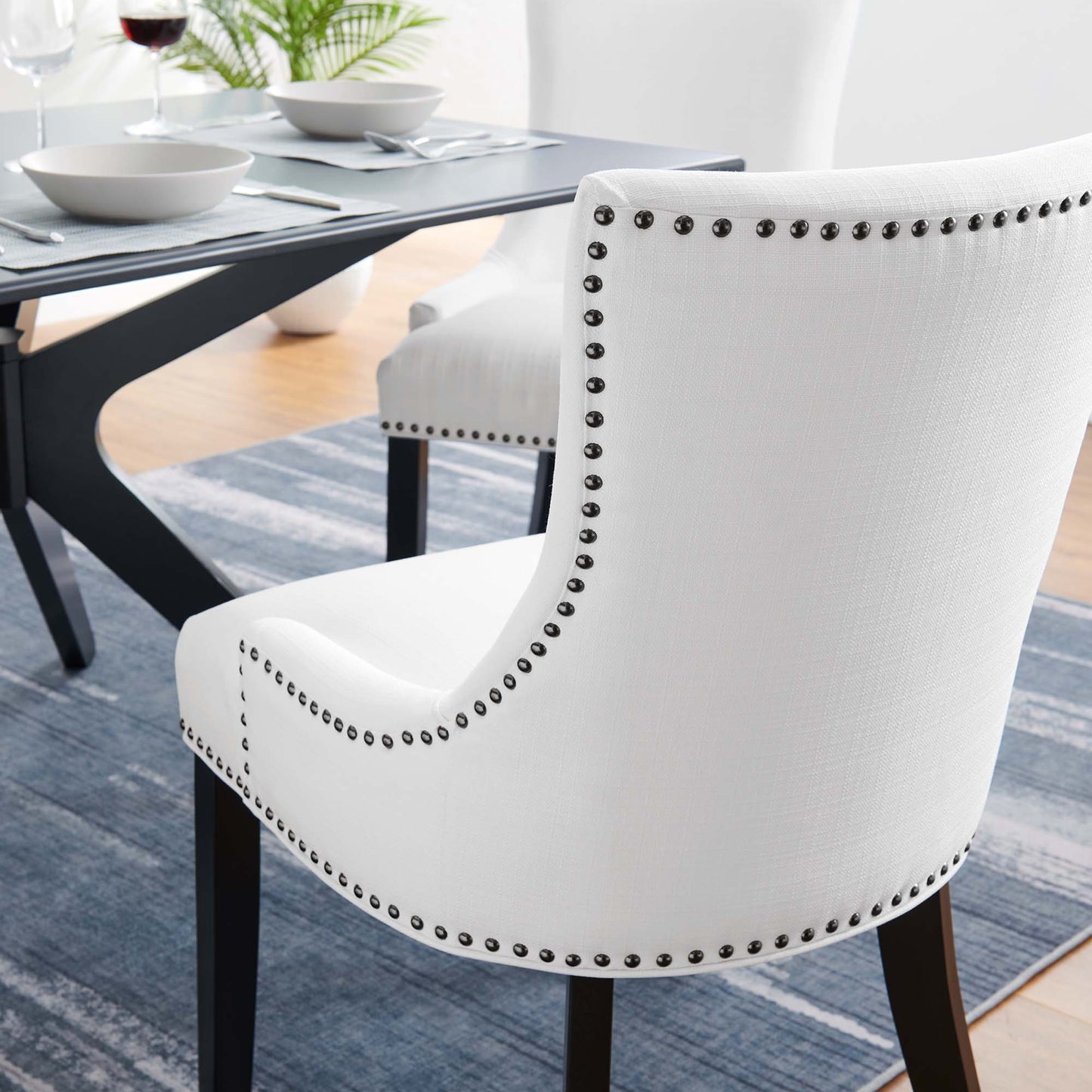 White upholstered dining chair with curved back, dark wood legs, silver nailhead trim, in a contemporary dining room.