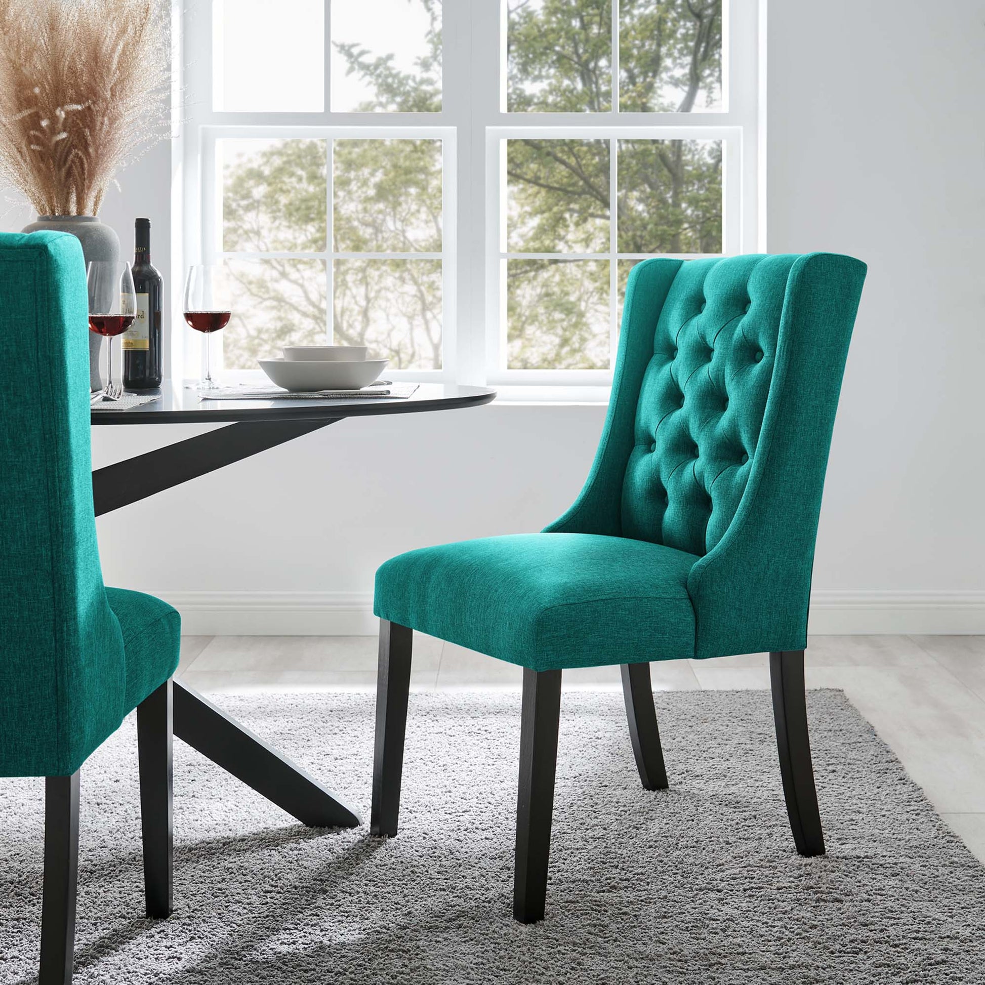 Teal fabric button-tufted dining chair with dark wooden legs around a round glass-top table in a bright modern dining room.