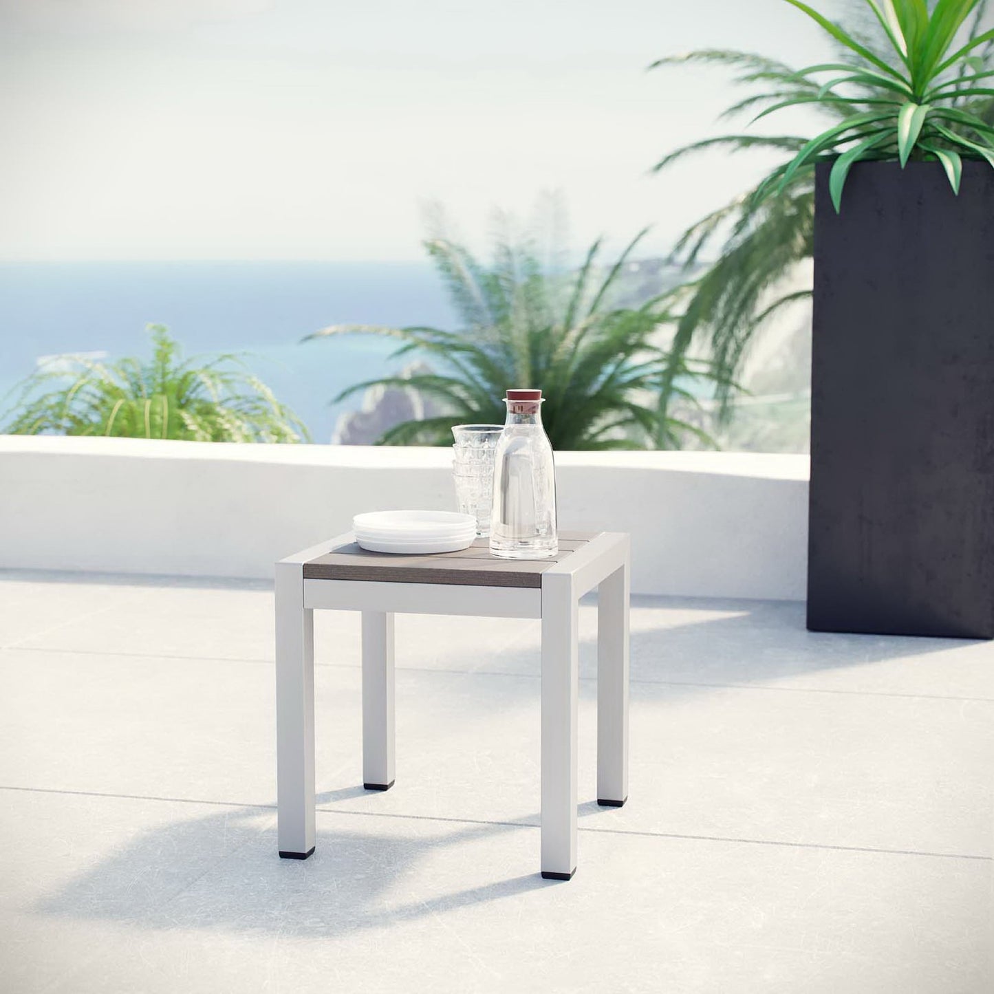 Gray outdoor aluminum side table on a light tiled patio with a glass water bottle and a white plate, sea view, modern minimalist.