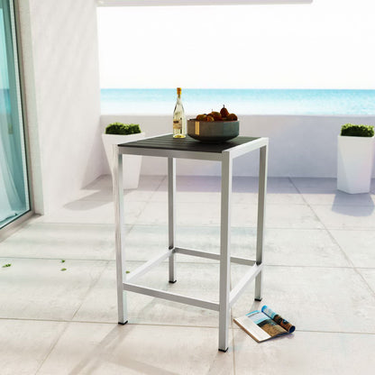 Silver-gray outdoor square bar table with gray legs, bottle and fruit bowl on top; magazine on tiled patio by the sea.