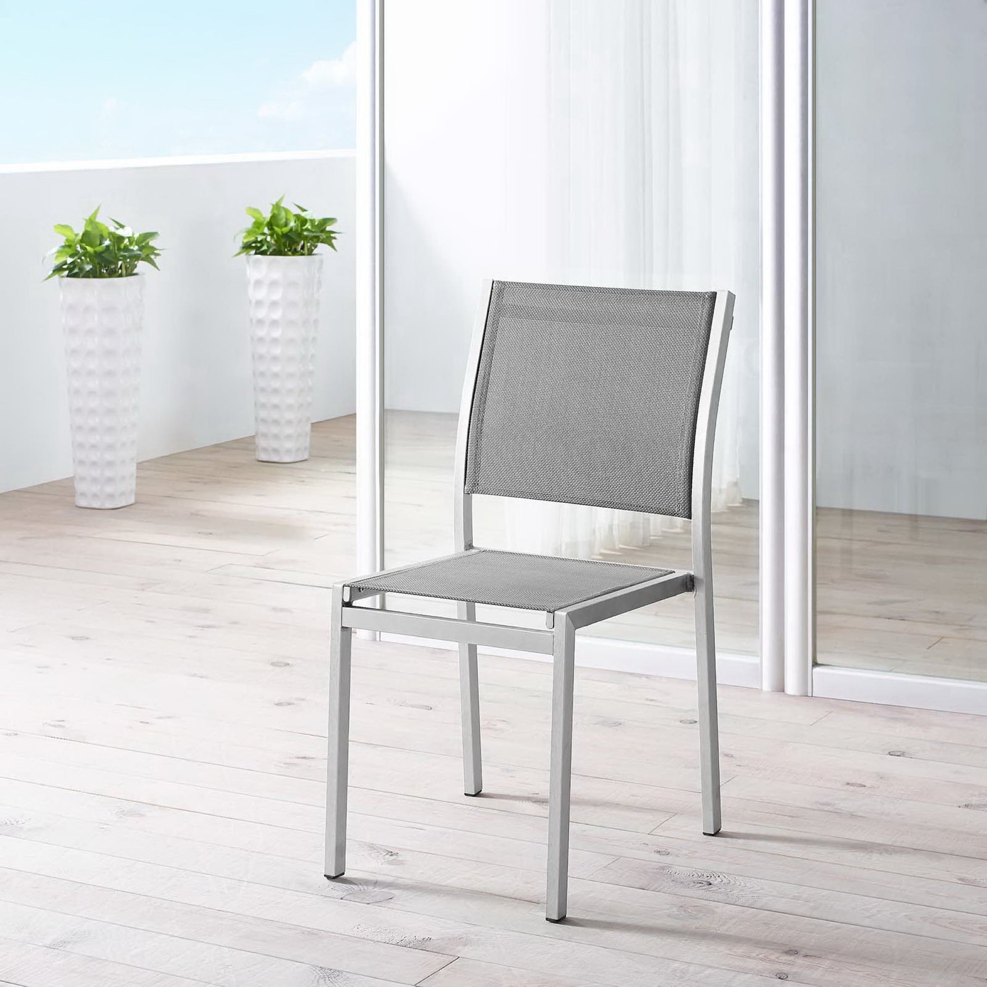 Silver-gray aluminum outdoor side chair with mesh seat and back on a light wooden deck, with glass doors and tall white planters.
