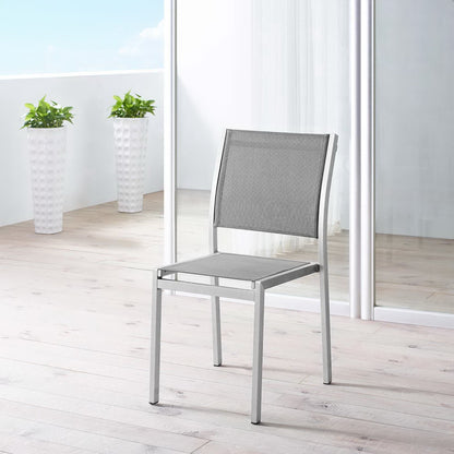 Silver-gray aluminum outdoor side chair with mesh seat and back on a light wooden deck, with glass doors and tall white planters.