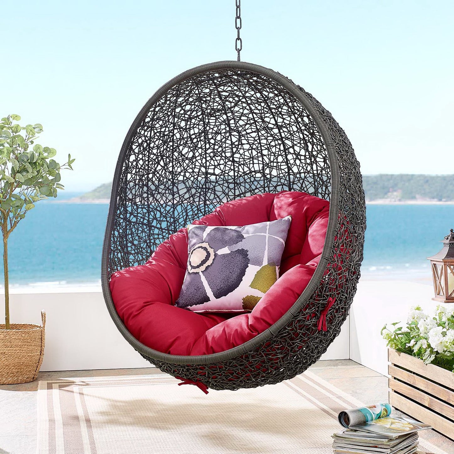 Outdoor patio swing chair with a stand, dark gray frame and red cushions, floral pillow, on a seaside patio.