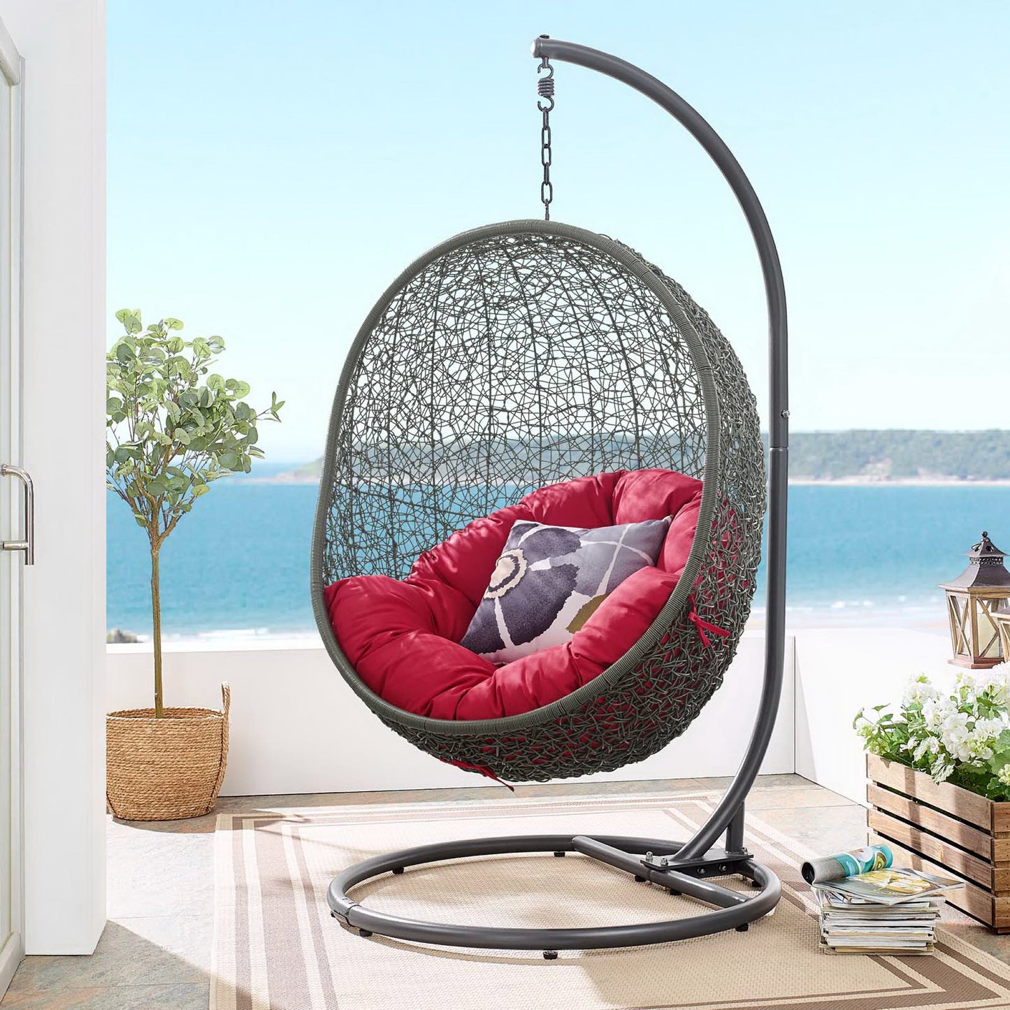 Egg-shaped wicker hanging chair with red cushions, suspended from a curved metal stand over a circular base, with sea view.