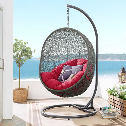 Egg-shaped wicker hanging chair with red cushions, suspended from a curved metal stand over a circular base, with sea view.
