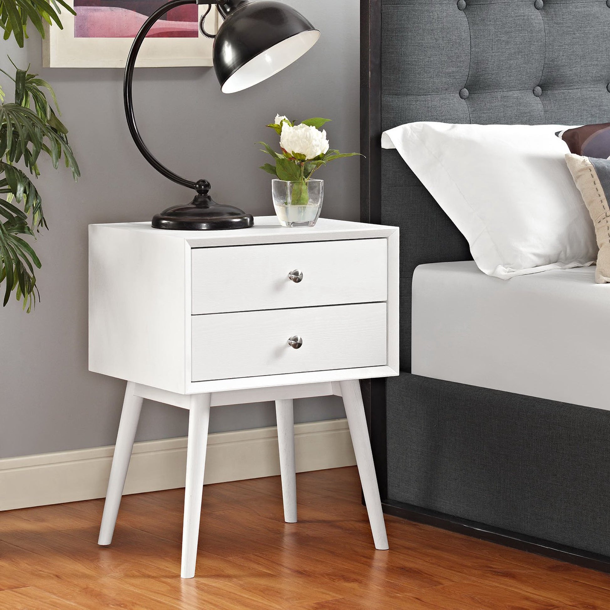 White mid-century nightstand with two drawers, tapered legs, metal knobs, beside a grey tufted bed in a modern bedroom.