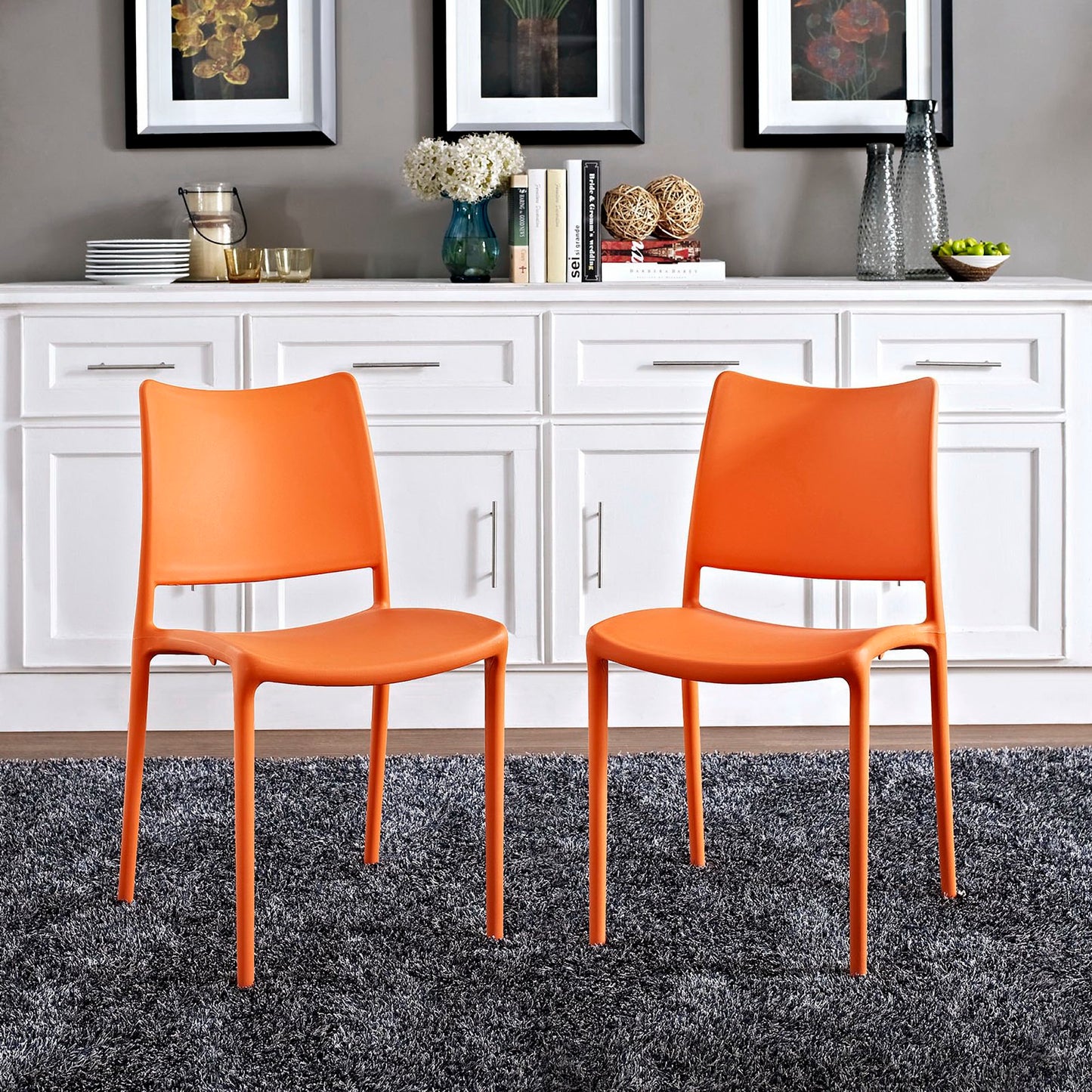 Two bright orange molded plastic dining chairs with slender metal legs on a gray rug in a modern kitchen with white cabinetry.
