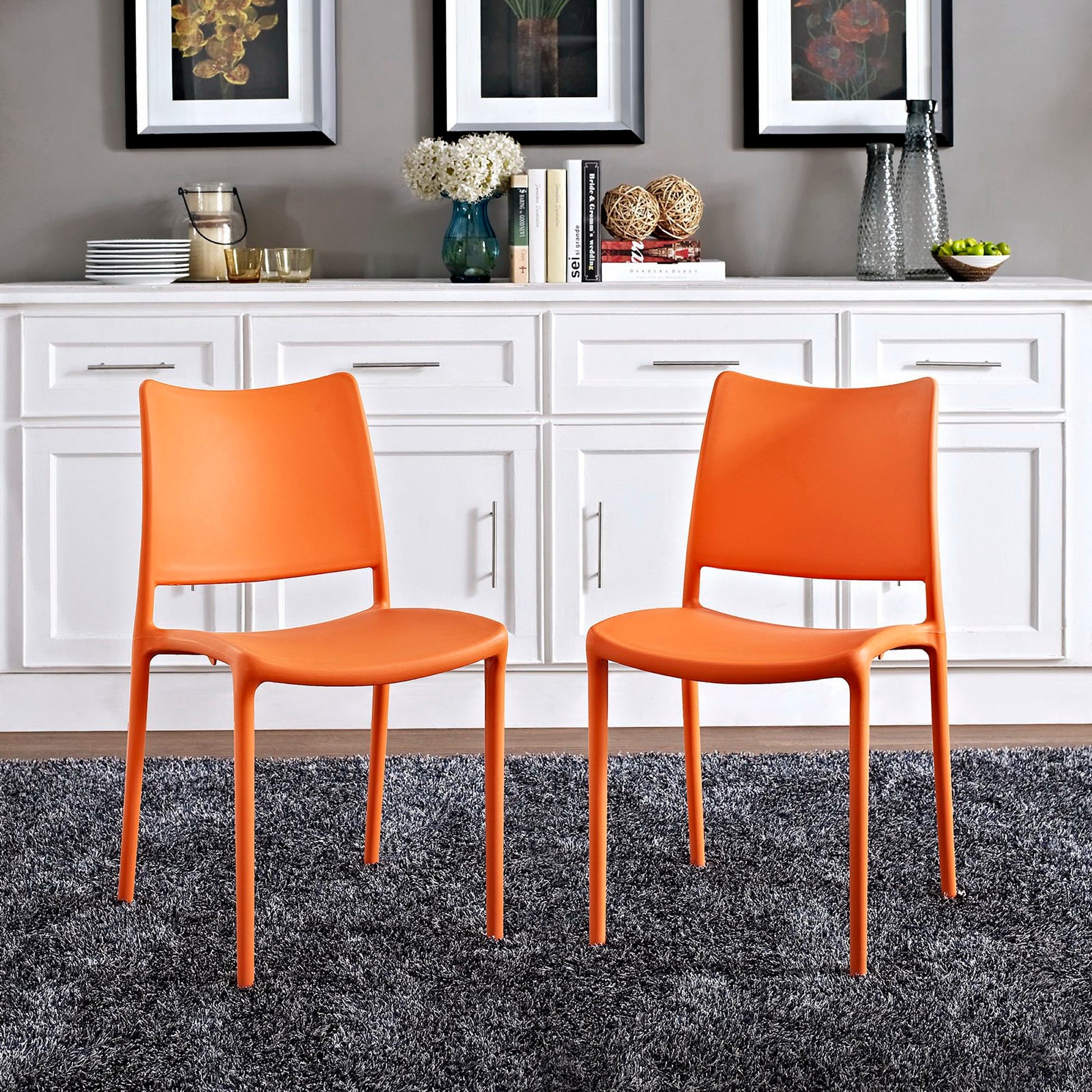 Two bright orange molded plastic dining chairs with slender metal legs on a gray rug in a modern kitchen with white cabinetry.