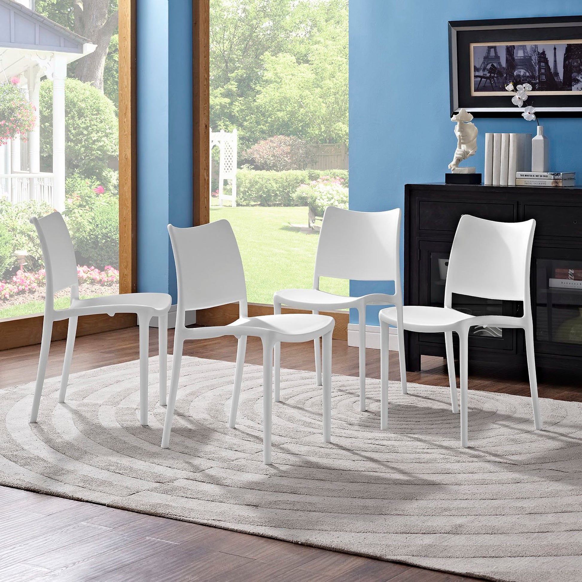 Four white modern plastic dining chairs around a circular rug in a sunlit room with blue walls and a black sideboard.