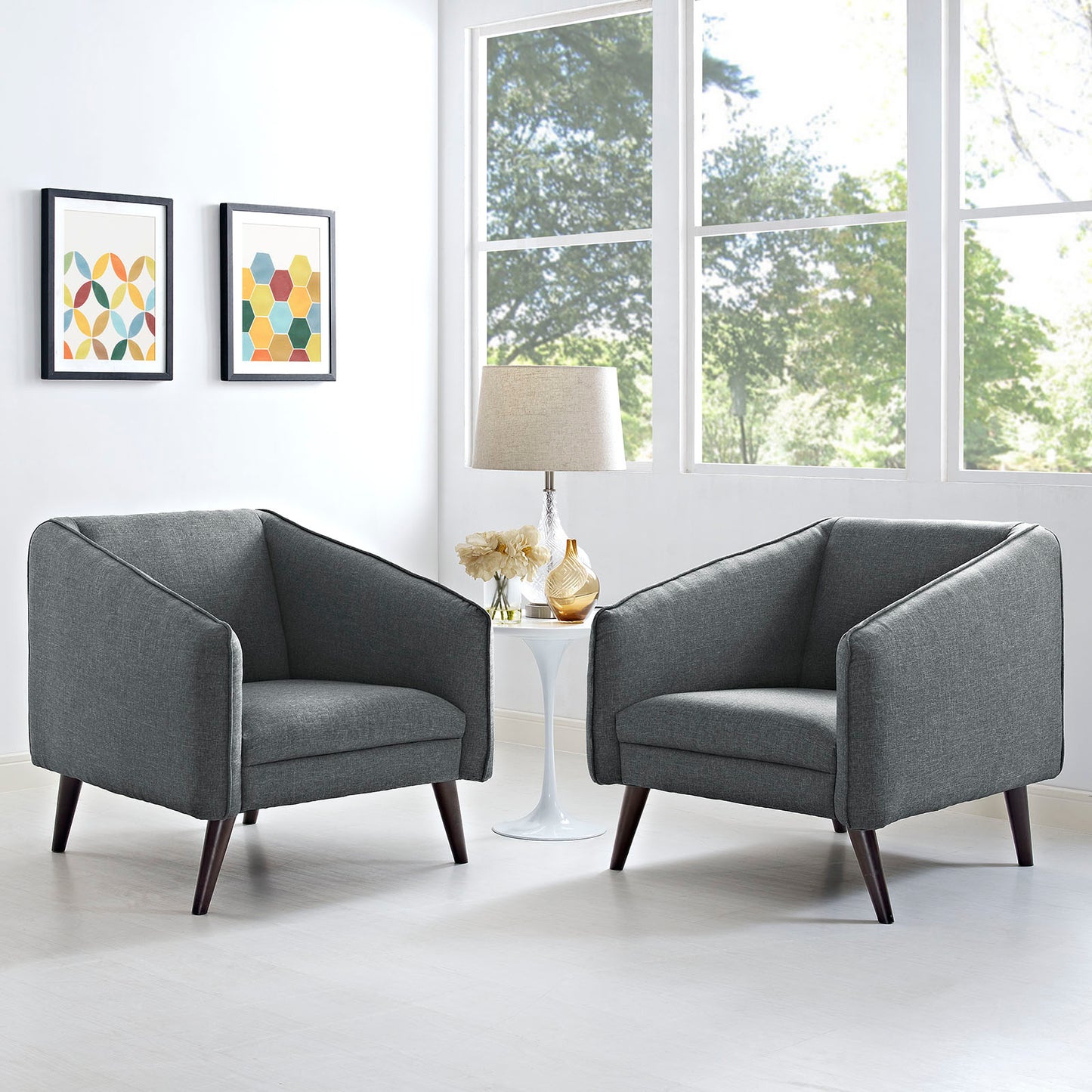 Two gray mid-century armchairs with dark legs flank a white round side table with lamp; bright room with geometric wall art.