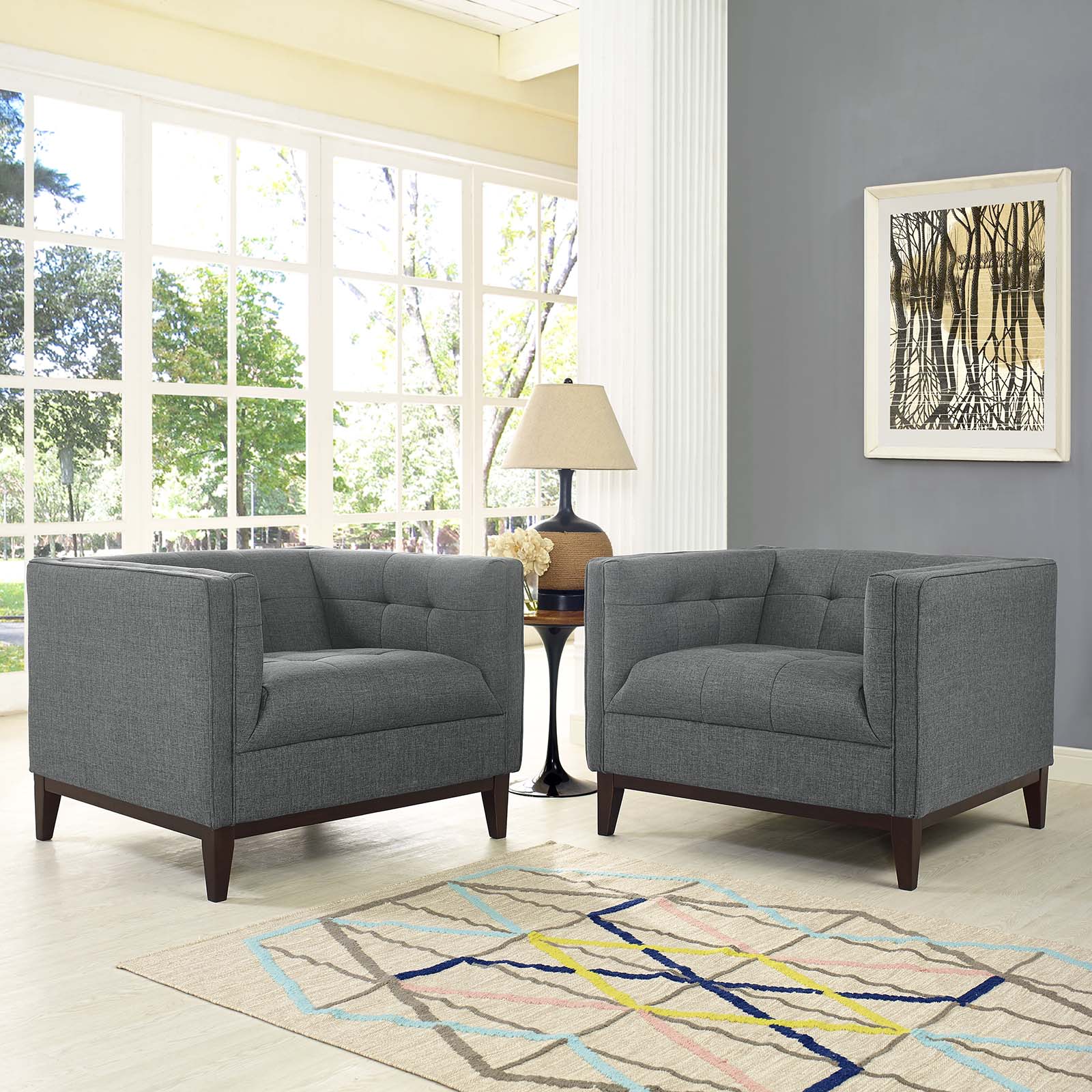 Two gray fabric armchairs with dark wood legs in a bright living room, between a round table and lamp; geometric rug, art.