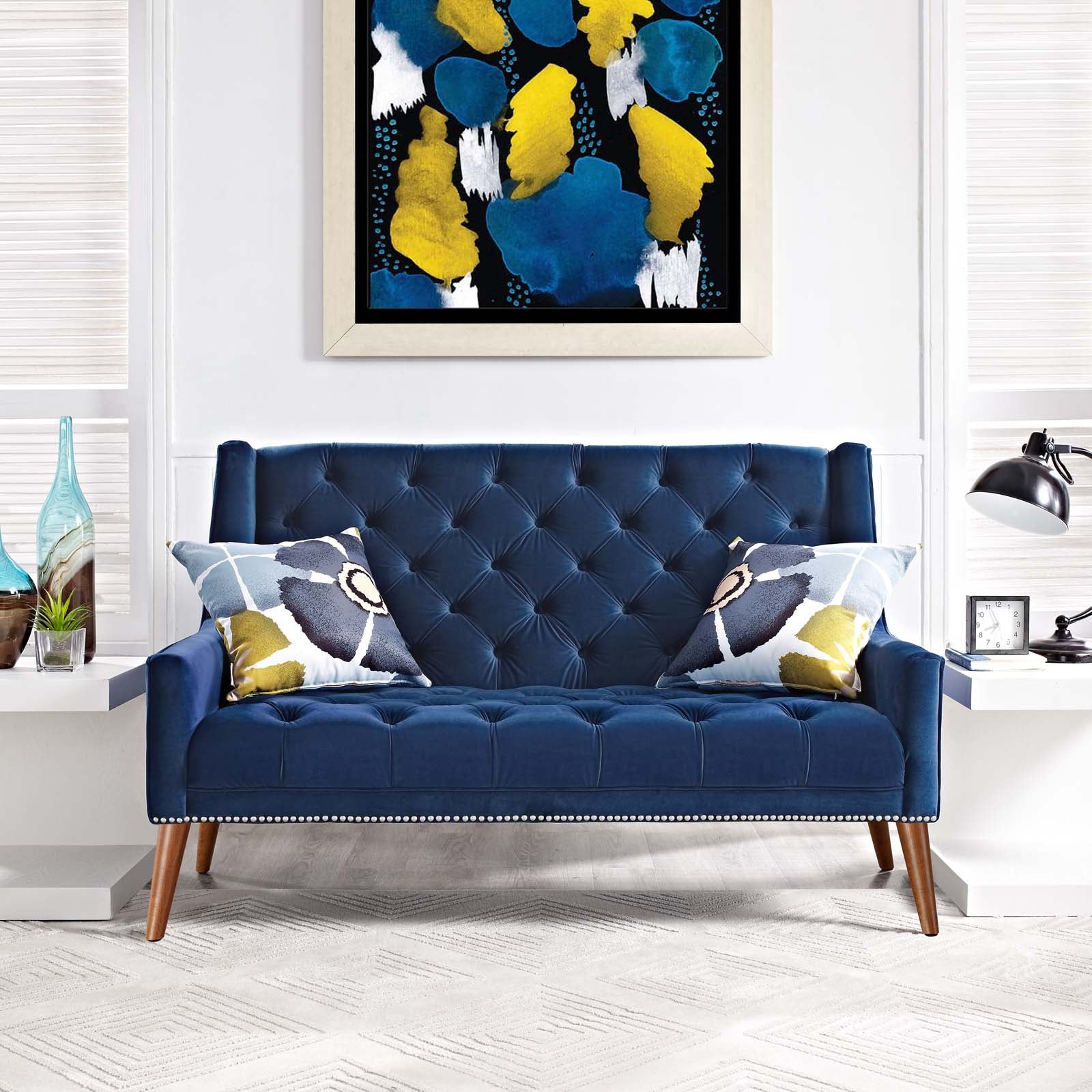 Navy tufted loveseat with nailhead trim and wooden legs, two geometric pillows; abstract blue-yellow art on white wall.