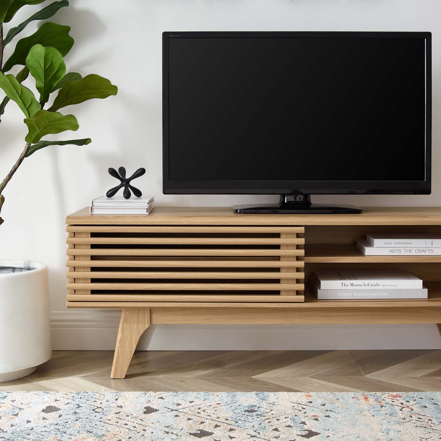 Oak TV console with slatted front, flat-screen TV on top, potted plant left, black sculpture, and books on shelf.