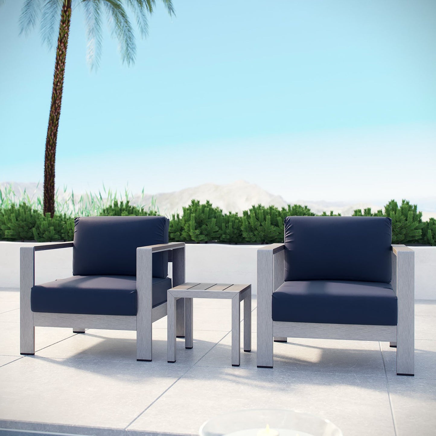Two navy cushions on light-gray outdoor lounge chairs with a small side table on a white patio, palm trees in background.