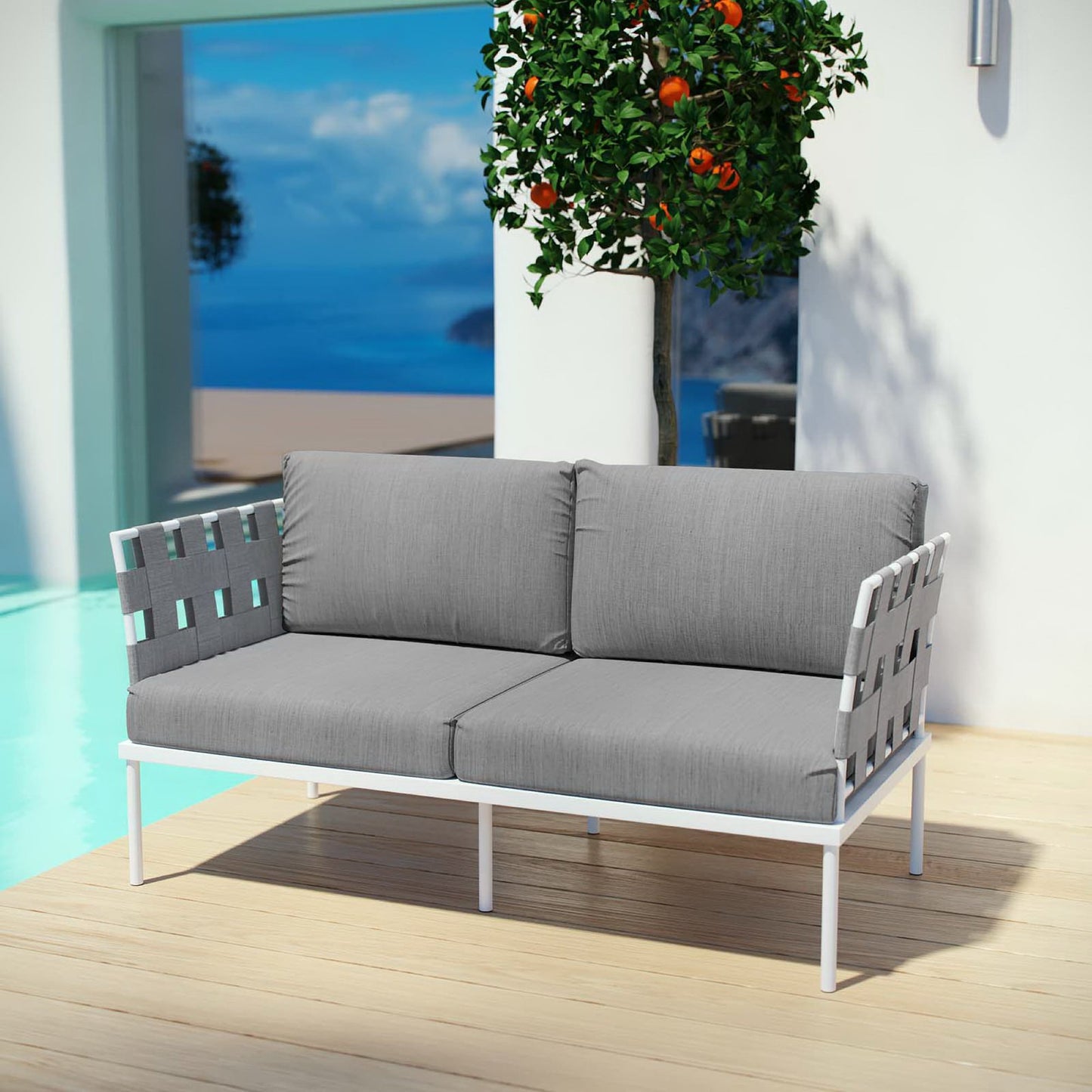 White outdoor loveseat with gray cushions and woven arms on a wooden deck beside a pool and orange tree.
