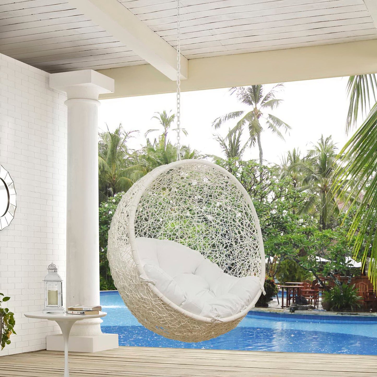White wicker hanging egg chair with plush cushions, suspended by a chain over a pool in a tropical patio.