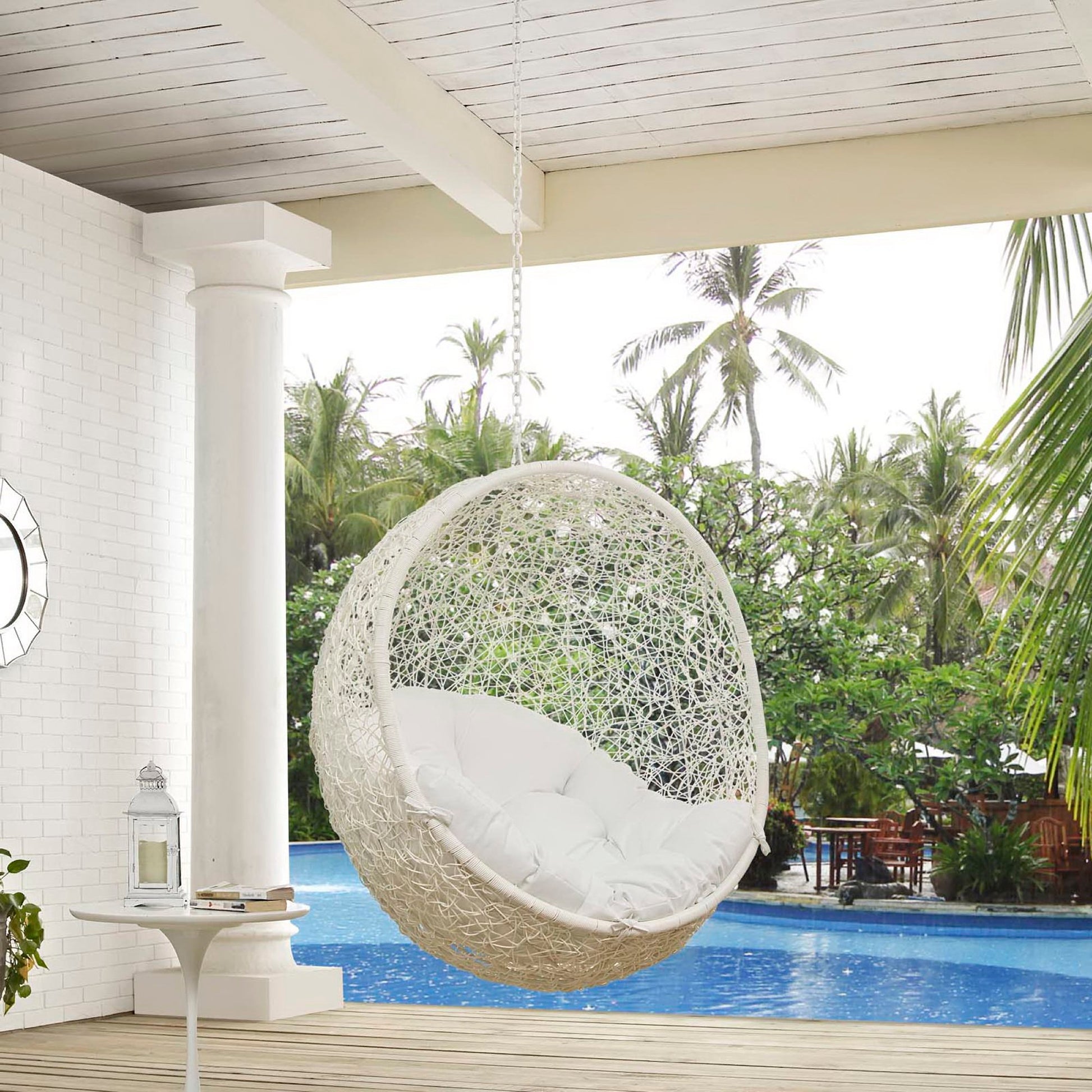 White wicker hanging egg chair with plush cushions, suspended by a chain over a pool in a tropical patio.