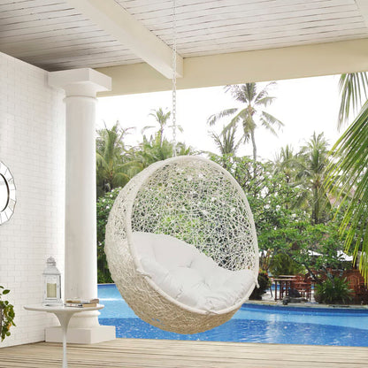 White wicker hanging egg chair with plush cushions, suspended by a chain over a pool in a tropical patio.