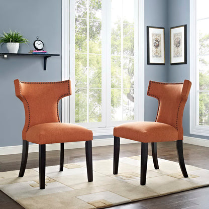 Pair of orange upholstered dining side chairs with nailhead trim, curved backs, and dark wood legs in a bright room with large windows.