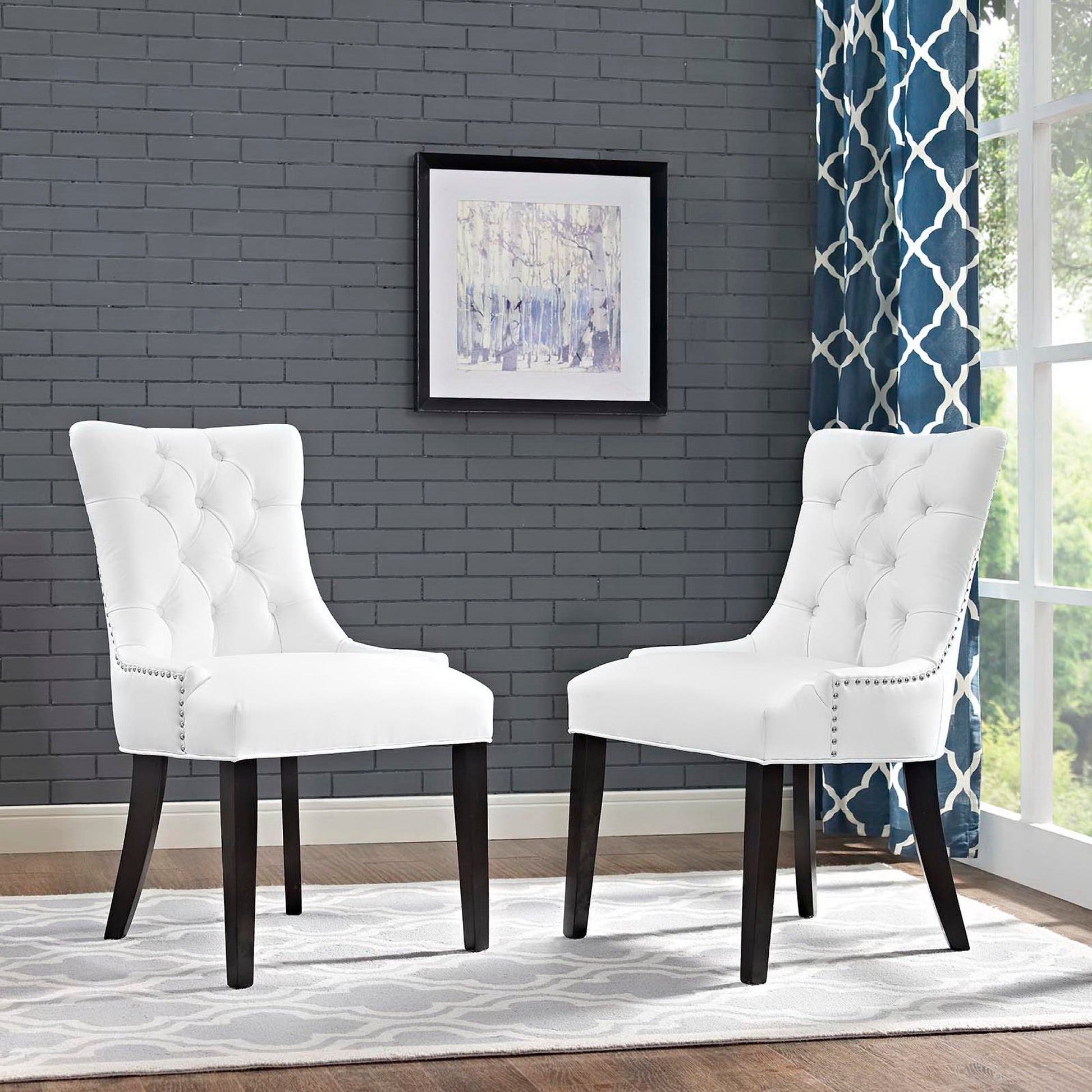 Pair of white tufted dining chairs with dark wood legs and nailhead trim in a gray brick room with blue patterned curtains.