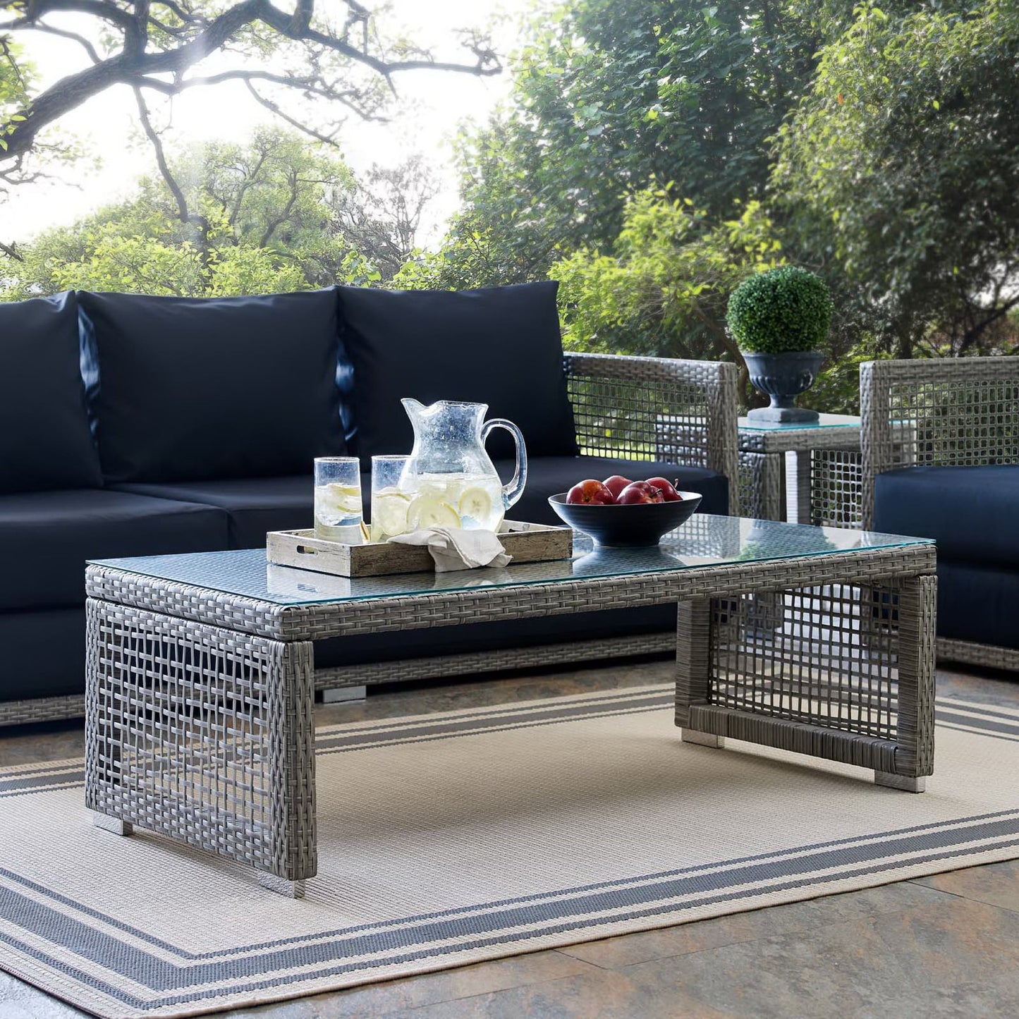 Gray rattan outdoor coffee table with glass top; tray with pitcher and glasses and a bowl of apples.