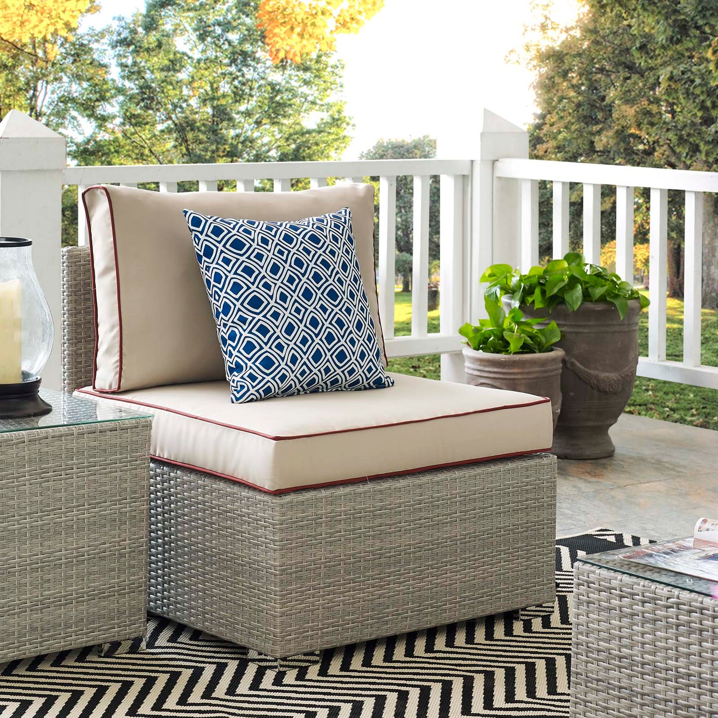Outdoor beige wicker lounge chair with thick cushion and blue geometric throw pillow on a chevron rug by planters.