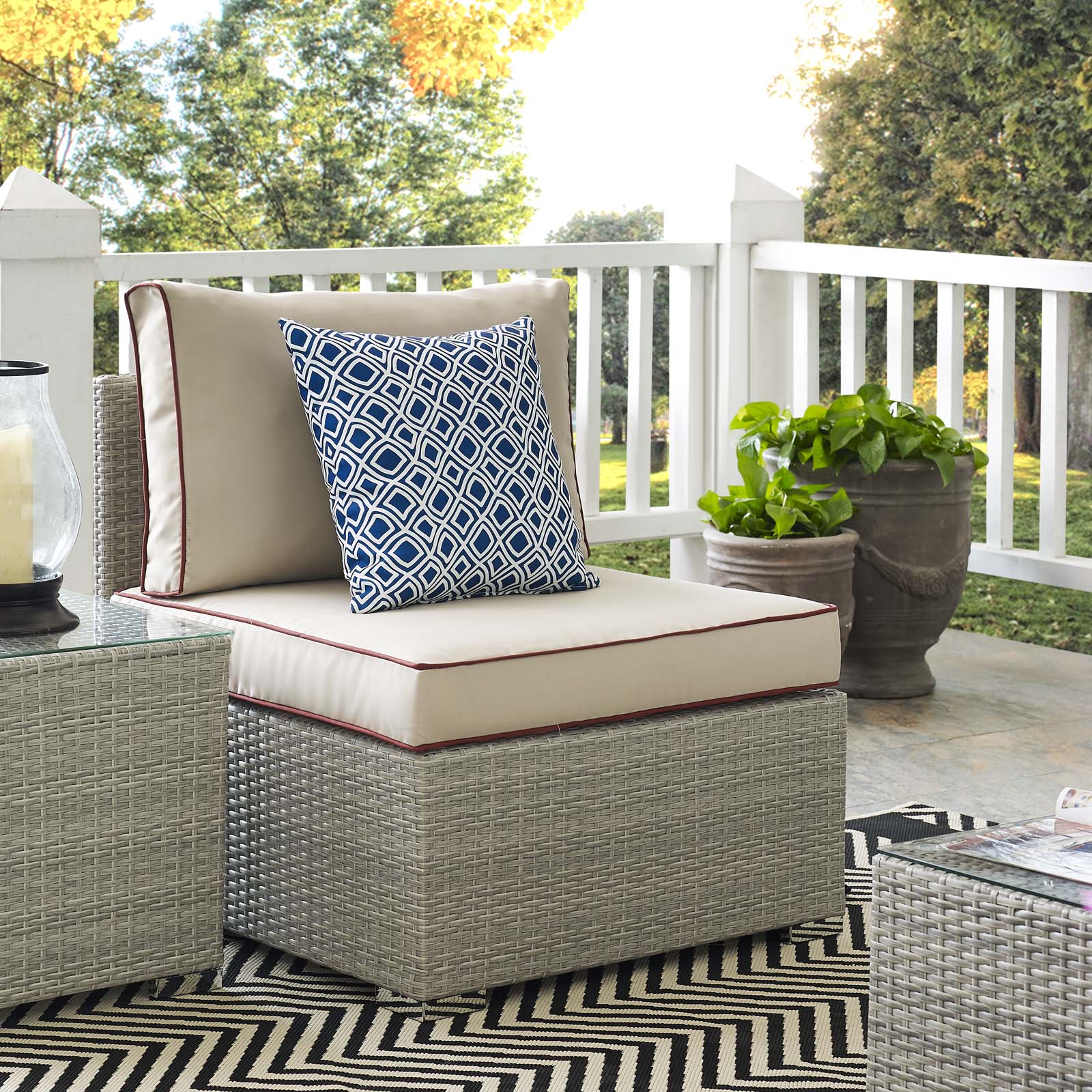 Outdoor beige wicker lounge chair with thick cushion and blue geometric throw pillow on a chevron rug by planters.