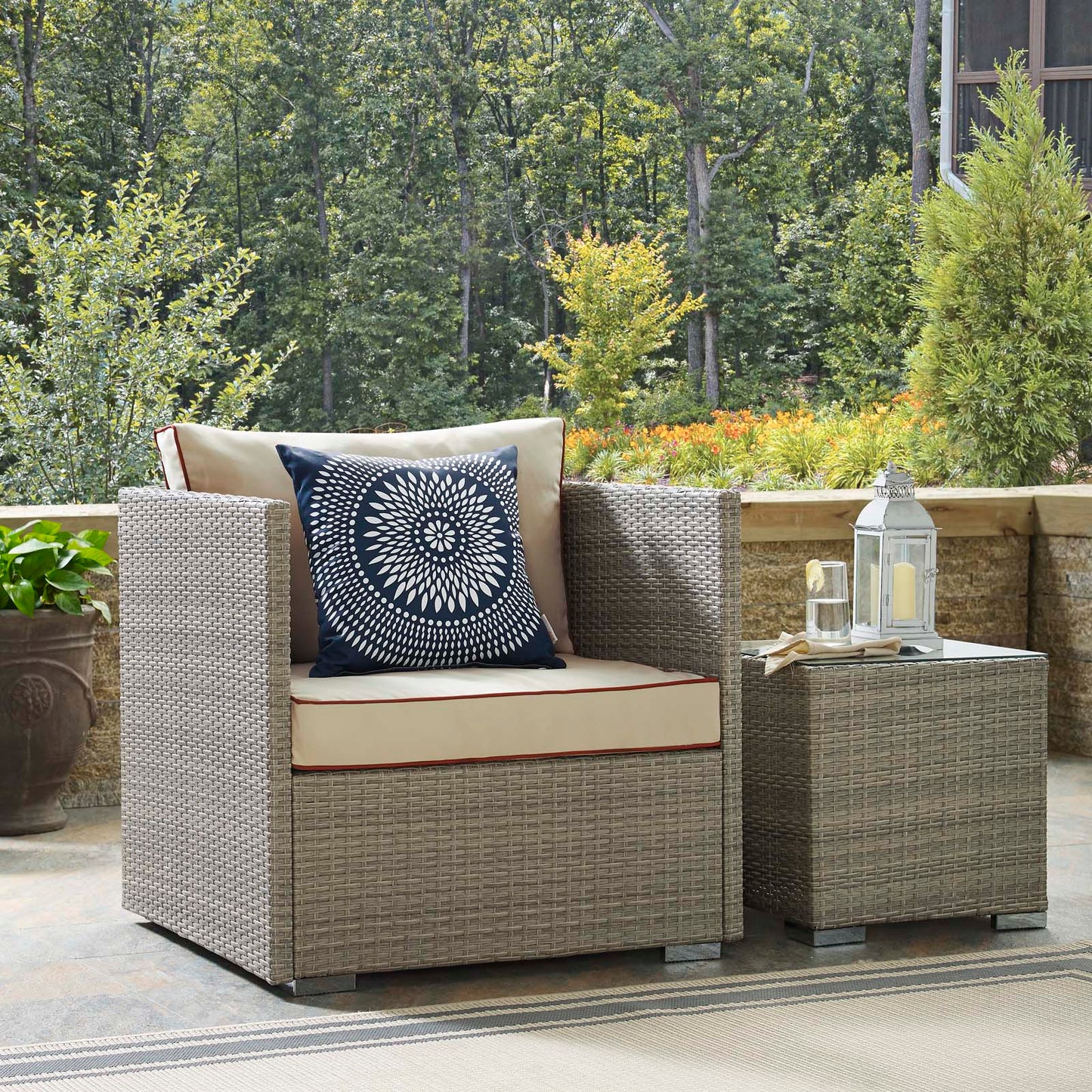 Beige woven outdoor rattan armchair with cream cushions, a navy mandala pillow, and a matching side table topped with lanterns.