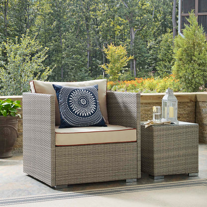 Beige woven outdoor rattan armchair with cream cushions, a navy mandala pillow, and a matching side table topped with lanterns.