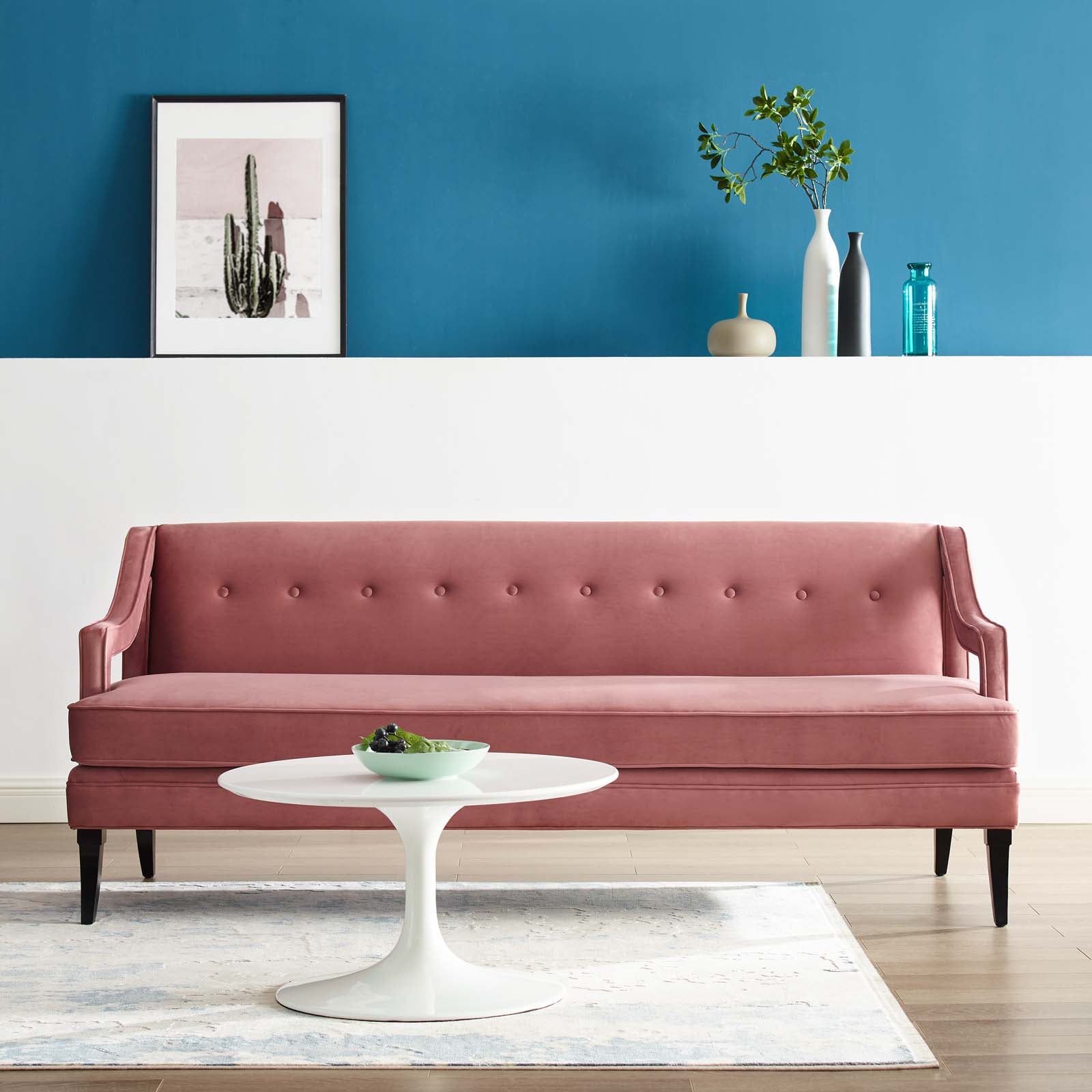 Dusty rose velvet tufted sofa with dark legs in a chic living room, white round coffee table, blue wall and cactus artwork.