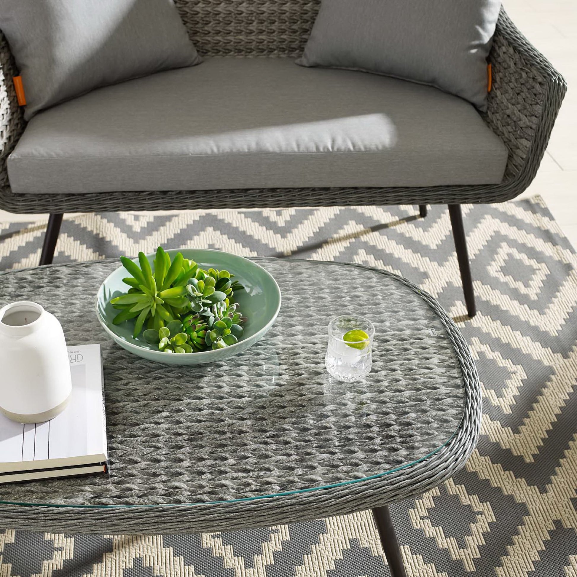Round glass-top wicker coffee table with a succulent bowl on a geometric gray-beige outdoor rug.